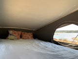 Inside an rv rental pop-top, a bed with pillows and a doll faces a scenic lake view through a mesh window. Ideal for camping.