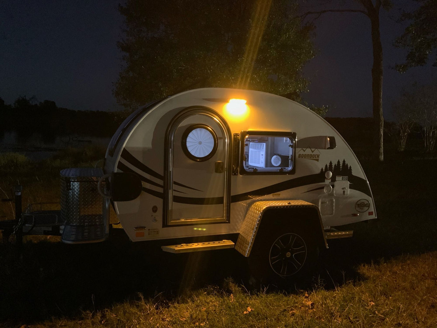 Cozy teardrop RV rental illuminated at a dark, serene waterside campsite. Perfect for night camping adventures.