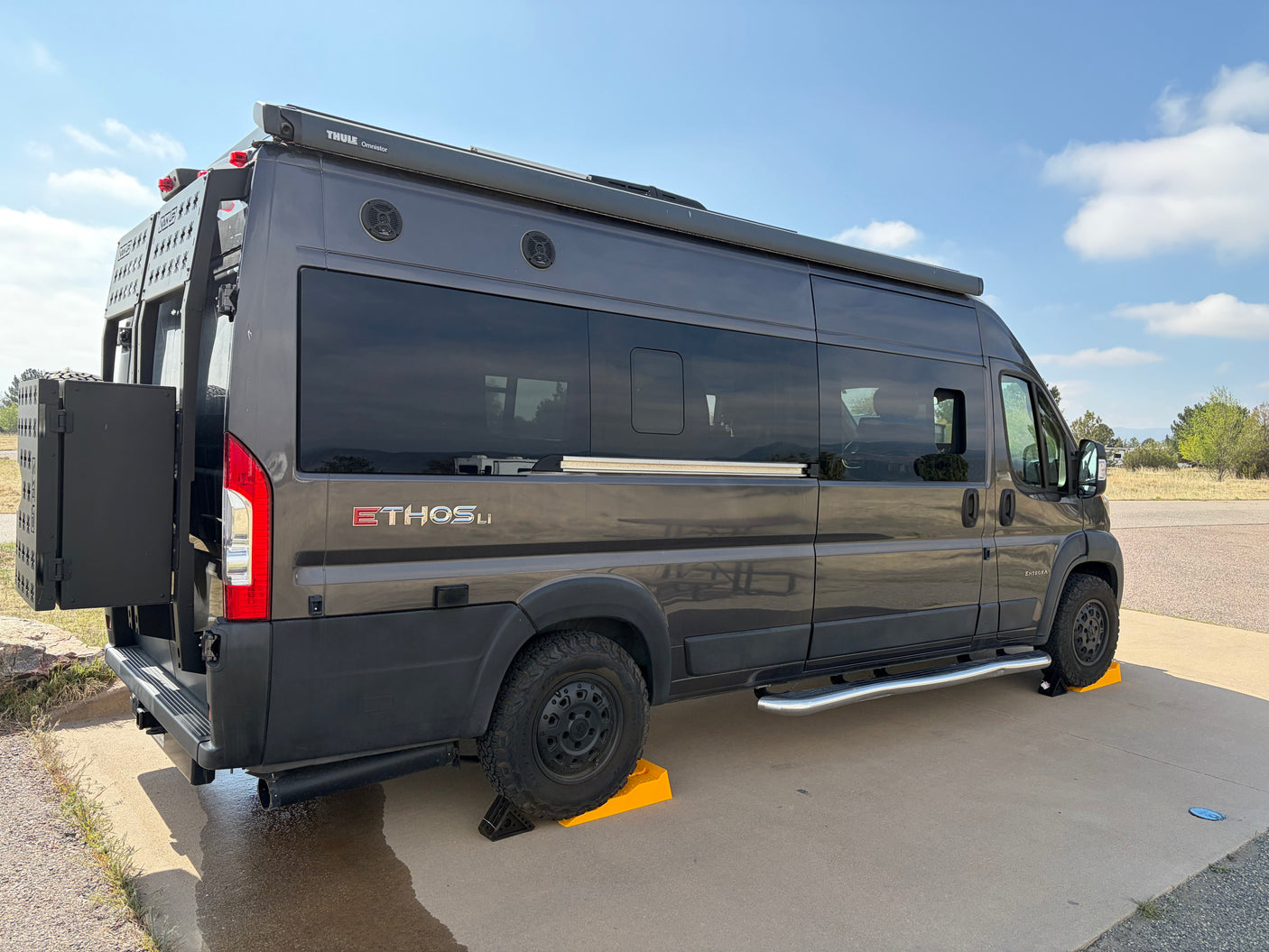 This dark gray Ethos Li camper van rv rental is parked with yellow leveling blocks. It features a Thule awning and rear cargo.