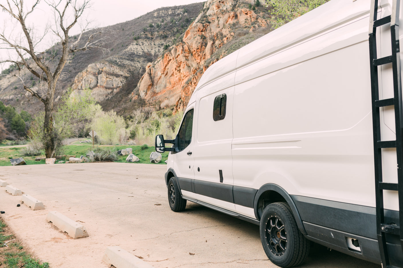 White rv rental camper van with a ladder, parked near rugged mountains and spring trees.