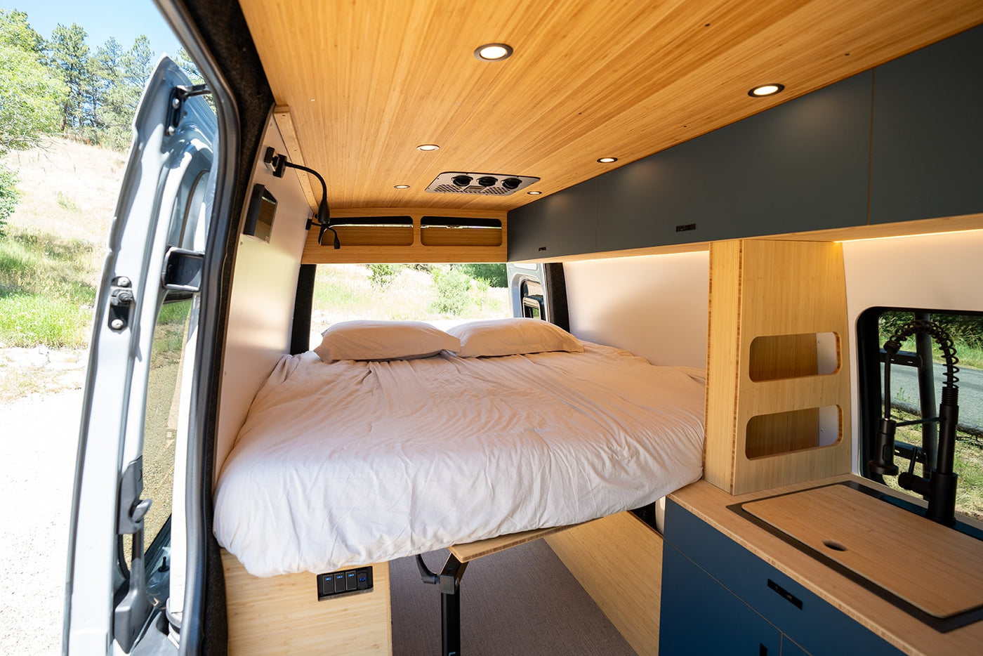 Cozy rv rental van interior: a large bed, sleek bamboo ceiling with lights, and a compact kitchen. Nature visible outside. — photo 7