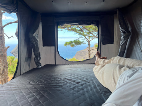 Relaxing in an RV rental's cozy rooftop tent, offering stunning ocean and cliff views. A perfect nature escape.