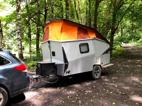 RV rental: A grey car tows a pop-up camper in a lush forest, parked on a leafy dirt path near a gate. Perfect for outdoor travel.
