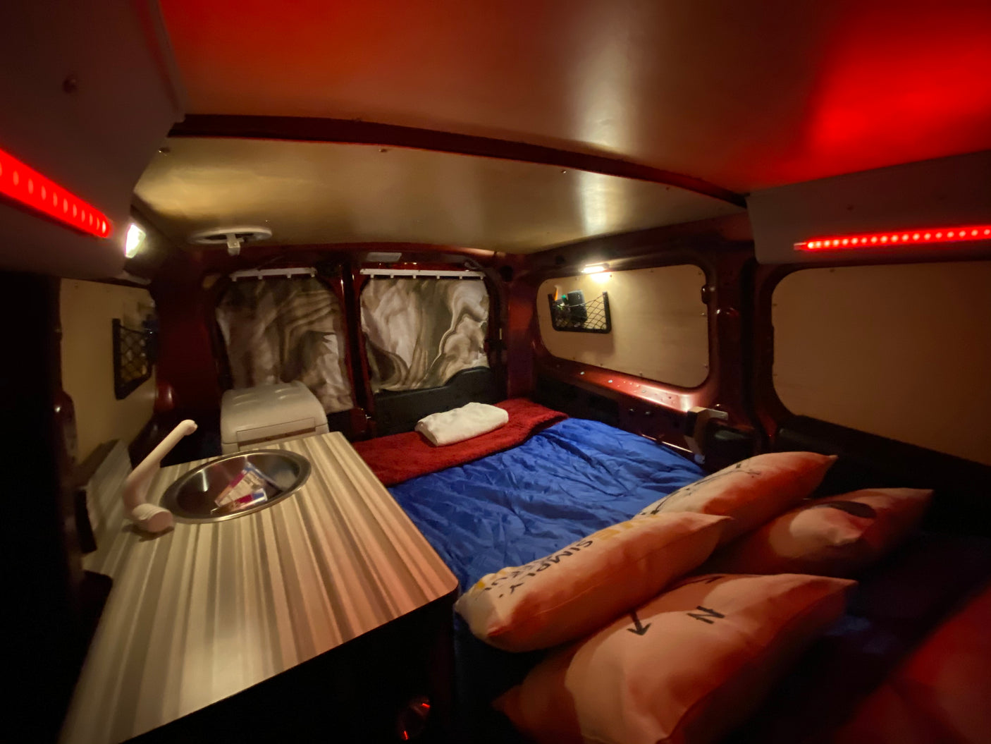 Cozy camper van interior, perfect for an RV rental, featuring a comfortable bed, compact kitchen with sink, and ambient red and white lighting.