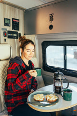 A woman in a cozy plaid shirt sips coffee and eats banana toast inside an RV rental, ready for travel adventures.