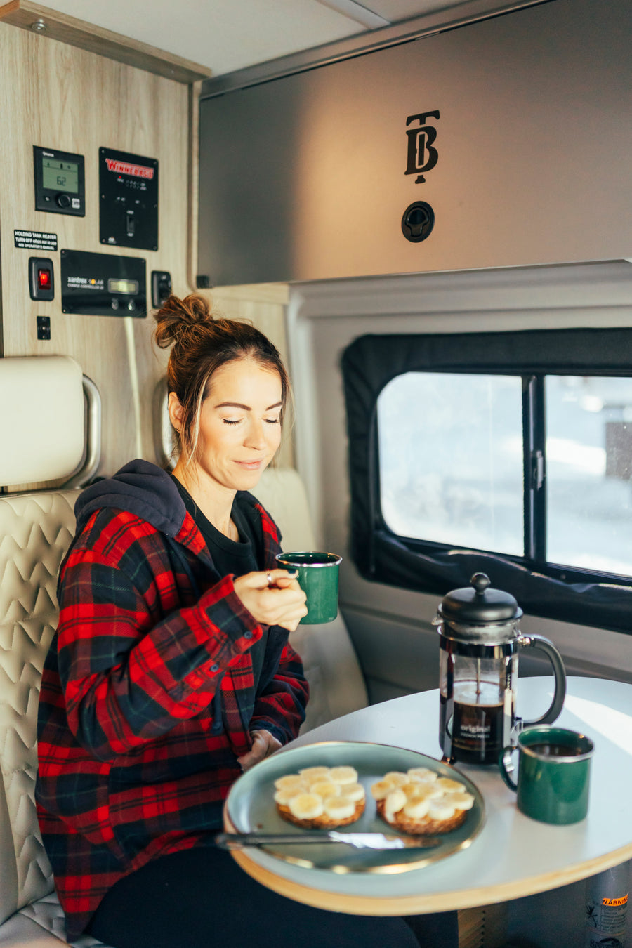 A woman in a cozy plaid shirt sips coffee and eats banana toast inside an RV rental, ready for travel adventures.