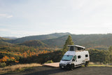 White RV rental camper van with a pop-top, parked at a scenic overlook, surrounded by vibrant autumn mountains.