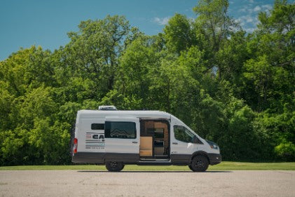 White camper van with open side door showcasing its interior, parked outdoors. Ideal for an RV rental trip surrounded by nature.