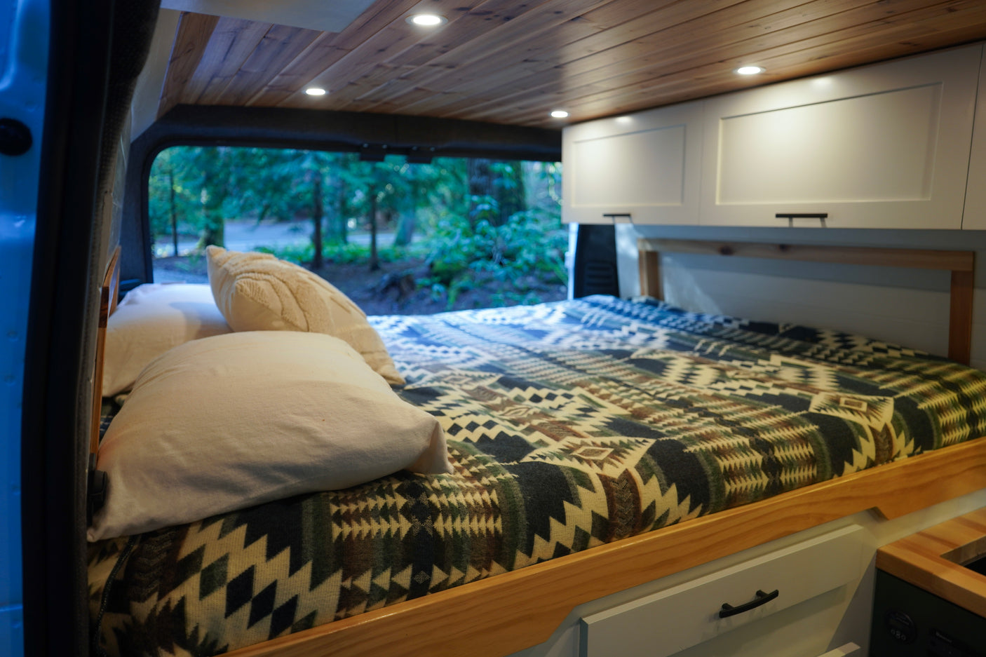 Cozy RV rental interior with a bed, wooden ceiling, cabinets, and a scenic forest view through the back window.
