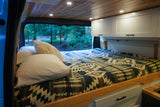 Cozy RV rental interior with a bed, wooden ceiling, cabinets, and a scenic forest view through the back window.