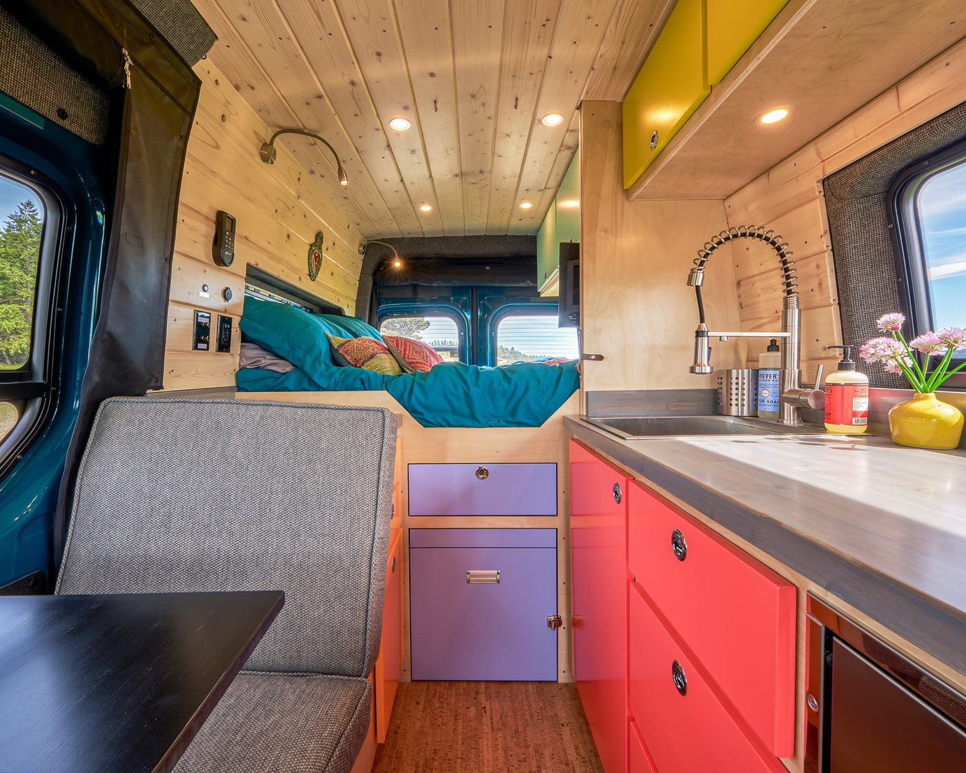 Vibrant RV rental interior: teal bed, bright kitchen with red and purple cabinets, and gray seating. Ready for adventure. — photo 7