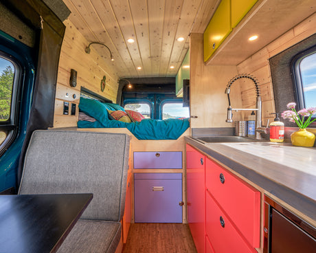 Vibrant RV rental interior: teal bed, bright kitchen with red and purple cabinets, and gray seating. Ready for adventure.