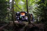 Adventurous rv rental: a camper van set up in a lush, mossy forest with open doors, illuminated interior, and drying wetsuits.