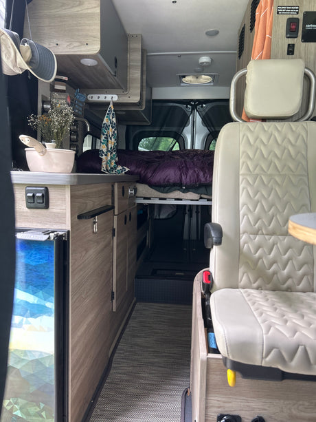 Step inside this cozy RV rental's interior, featuring a compact kitchen, comfortable bed, and front passenger seat.