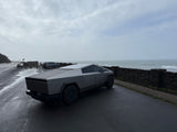 Sleek Tesla Cybertruck on a wet coastal road overlooking a stormy ocean. Forget an RV rental, this is modern exploration.