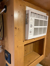 Compact Perfect Aire AC unit in a wooden RV rental interior, with a control panel and storage shelf below.