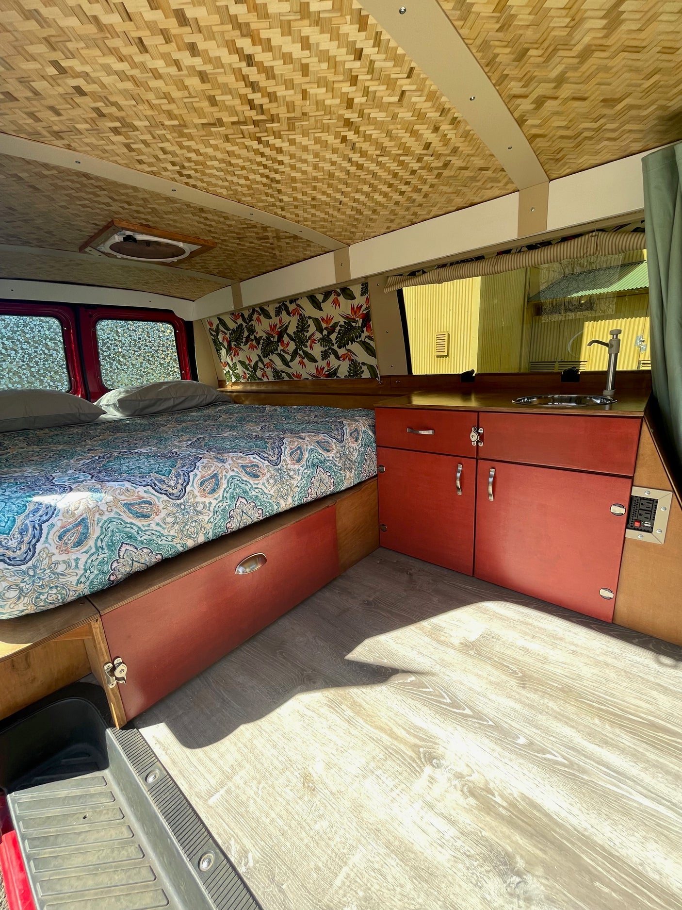 This RV rental's interior features a patterned bed, red storage cabinets, a small sink, and a unique woven ceiling.