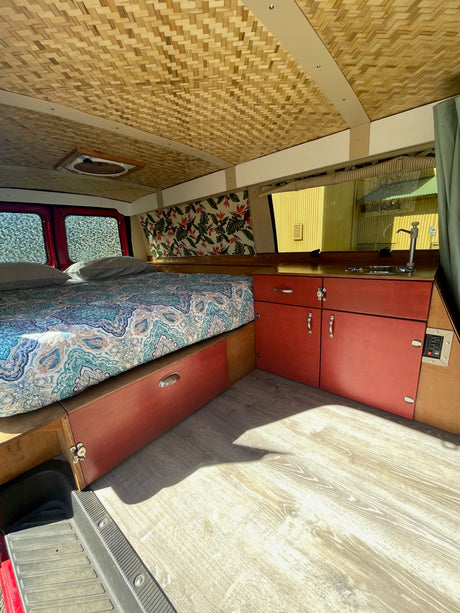 This RV rental's interior features a patterned bed, red storage cabinets, a small sink, and a unique woven ceiling.