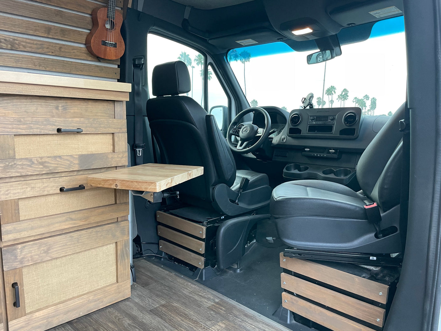 Explore this cozy RV rental camper van interior. Features include natural wood cabinets, a ukulele, and swivel seats overlooking palm trees.
