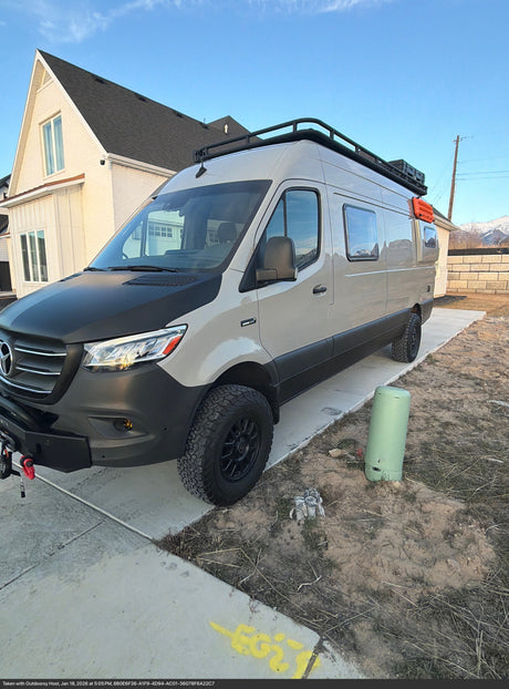 This tan & black Mercedes Sprinter rv rental, with a roof rack & off-road tires, is parked on a concrete pad next to a house.