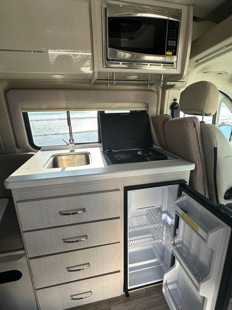 Inside this RV rental, a compact kitchen with sink, stovetop, microwave, and open fridge faces an ocean view.