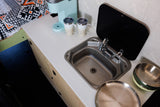 Explore this **rv rental's** compact kitchenette: sink, faucet, coffee maker, mugs, and bowls on a light countertop, next to cozy bedding.