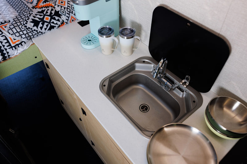 Explore this **rv rental's** compact kitchenette: sink, faucet, coffee maker, mugs, and bowls on a light countertop, next to cozy bedding. — photo 7