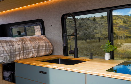 RV rental interior: A modern kitchen with a black sink, wood counter, and cooktop, alongside a plaid bed by scenic windows.