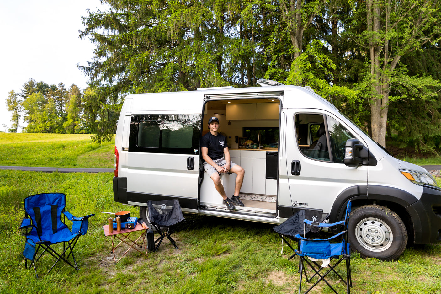 A man in his rv rental van sits by camping chairs, stove, and table, parked in a green, tree-lined field.