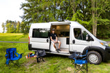 A man in his rv rental van sits by camping chairs, stove, and table, parked in a green, tree-lined field.
