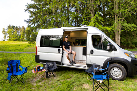 A man in his rv rental van sits by camping chairs, stove, and table, parked in a green, tree-lined field.