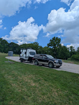 Black SUV towing a white No Boundaries travel trailer (RV rental) on a gravel lot near trees & a porta-potty, under a blue sky.