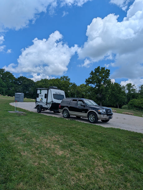 Black SUV towing a white No Boundaries travel trailer (RV rental) on a gravel lot near trees & a porta-potty, under a blue sky.