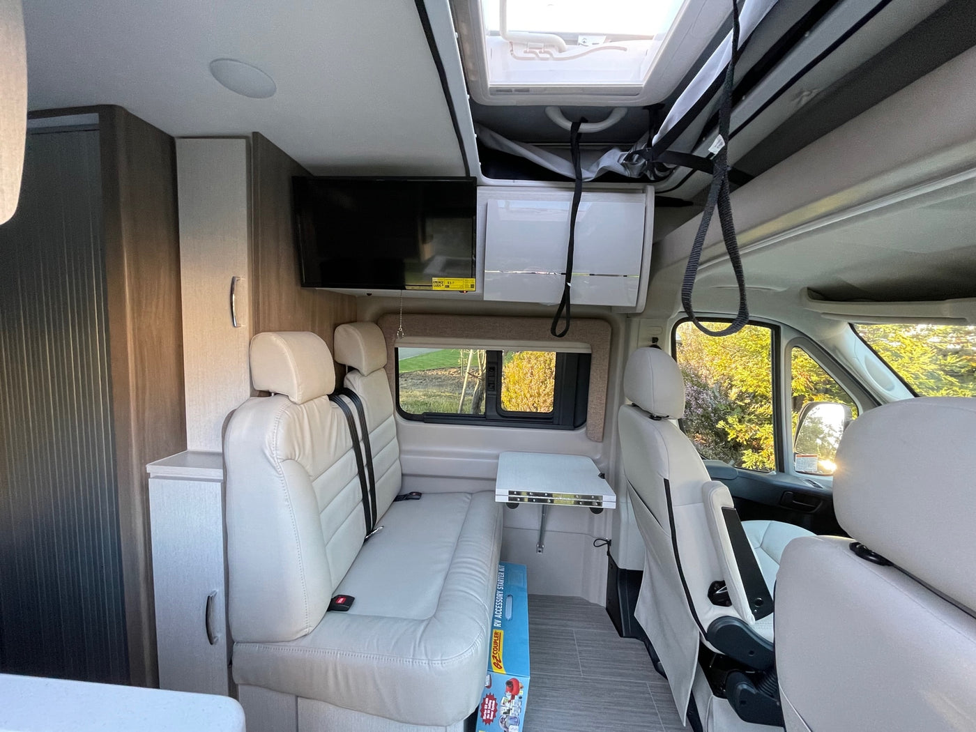 Inside a bright rv rental camper van, featuring light seating, an overhead TV, fold-out table, and skylight. Spacious interior. — photo 7