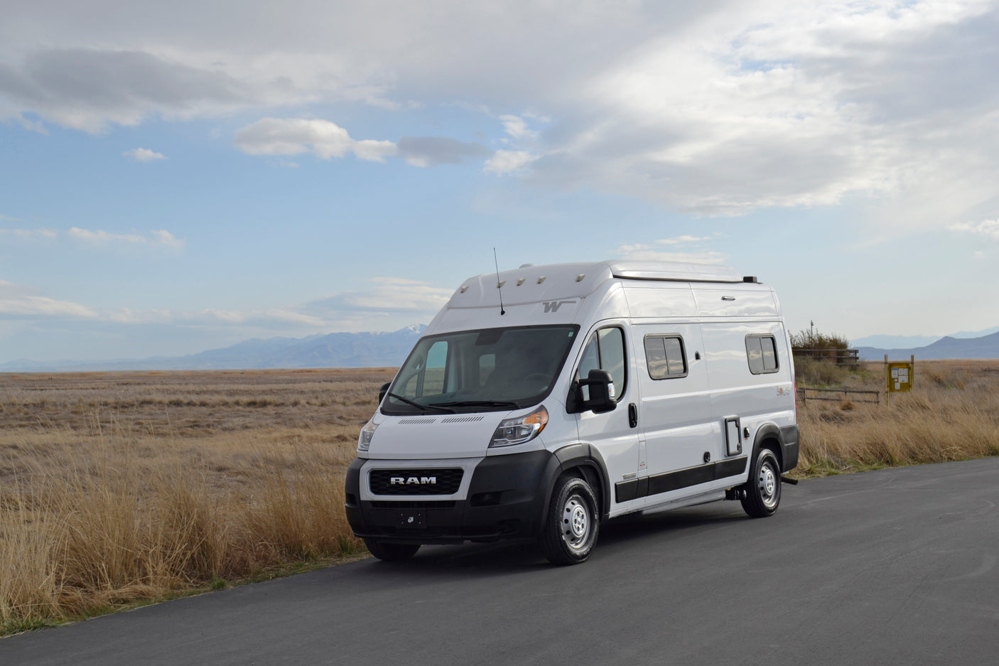This white RV rental van is parked on an asphalt road, set against a vast landscape of dry fields and distant mountains under a cloudy sky.