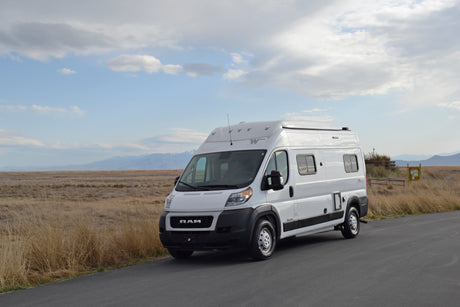 This white RV rental van is parked on an asphalt road, set against a vast landscape of dry fields and distant mountains under a cloudy sky.