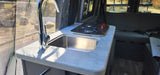 Compact kitchen in an RV rental, featuring a chrome faucet, stainless steel sink, and two-burner stove by a screened window.