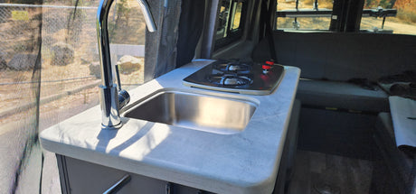 Compact kitchen in an RV rental, featuring a chrome faucet, stainless steel sink, and two-burner stove by a screened window.
