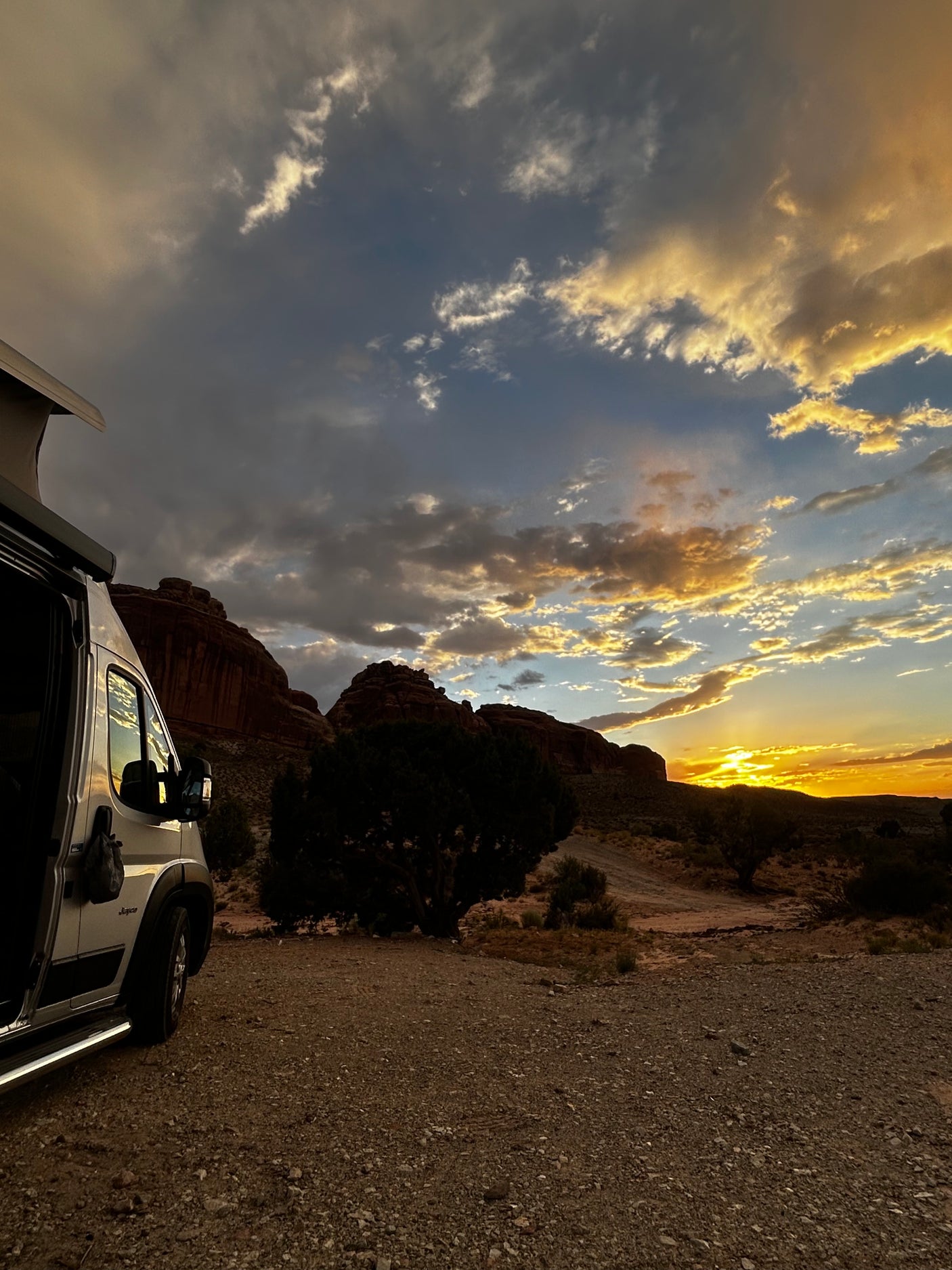 An RV rental is parked, capturing a breathtaking desert sunset over rock formations, with golden clouds filling the sky.