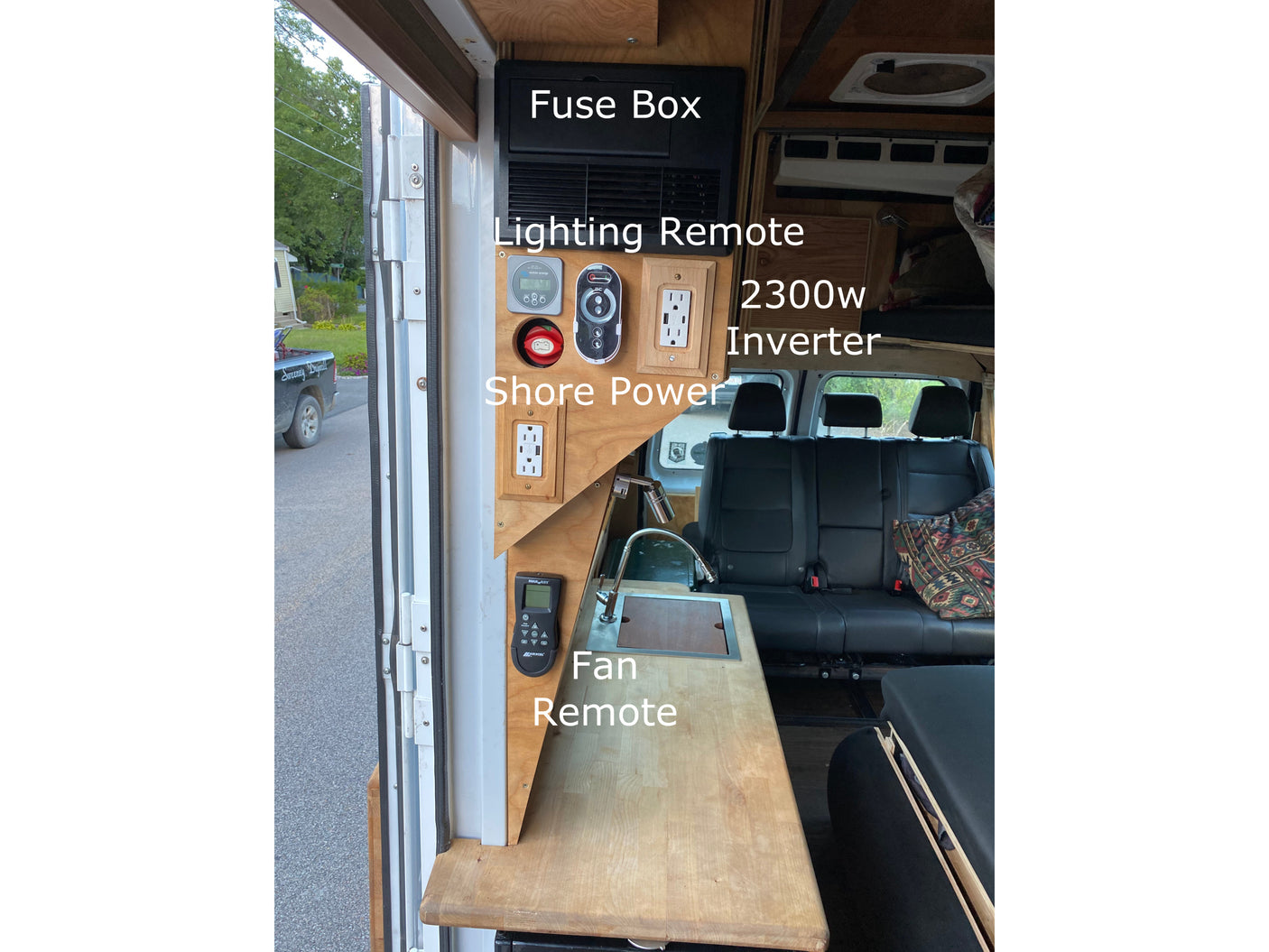 Inside an RV rental: a detailed electrical control panel featuring a fuse box, 2300w inverter, shore power outlet, lighting, and fan remotes, beside a sink.