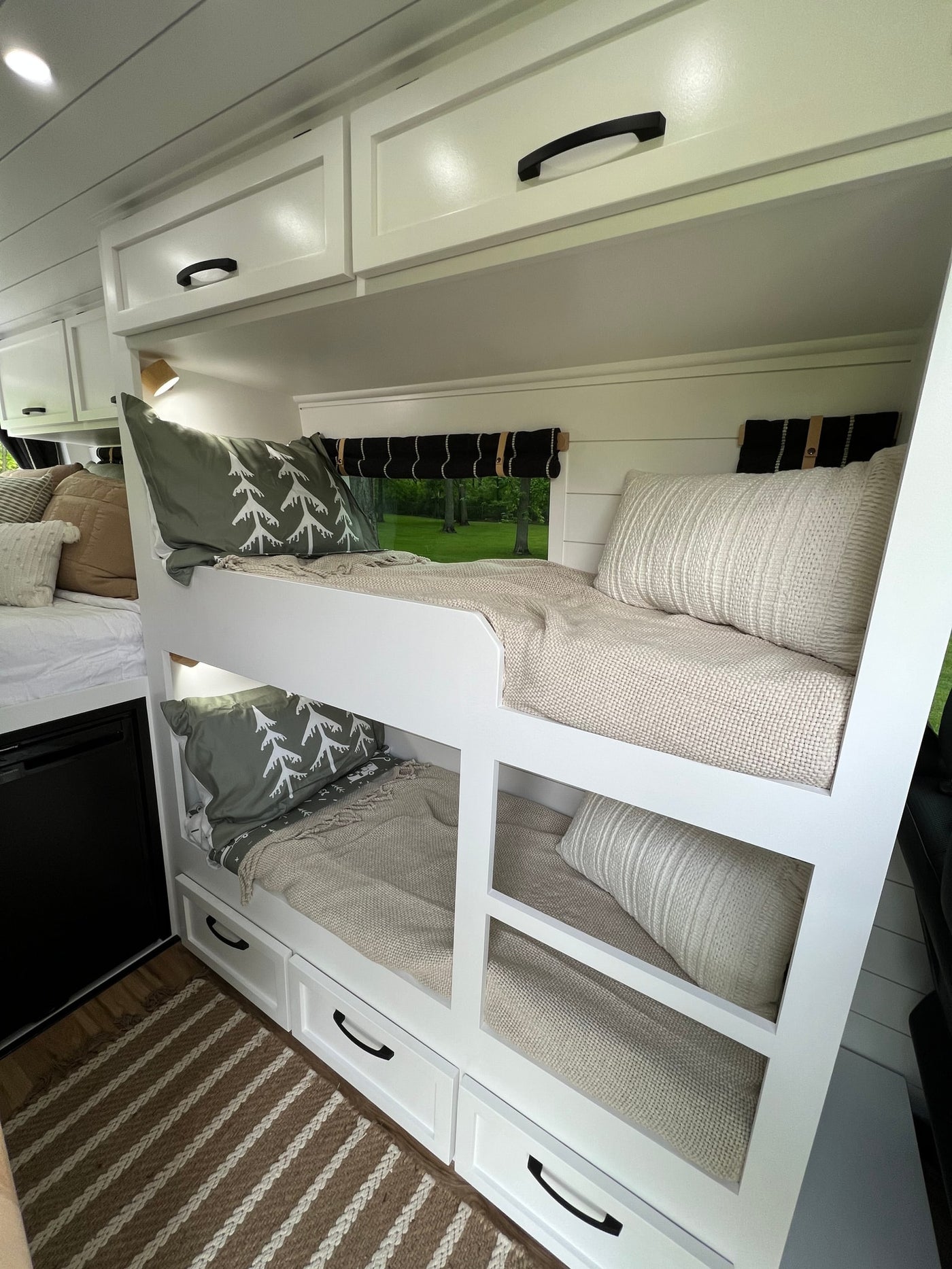 Inside a cozy **rv rental**: white bunk beds with drawers, plush pillows, and a window view of lush trees. — photo 12