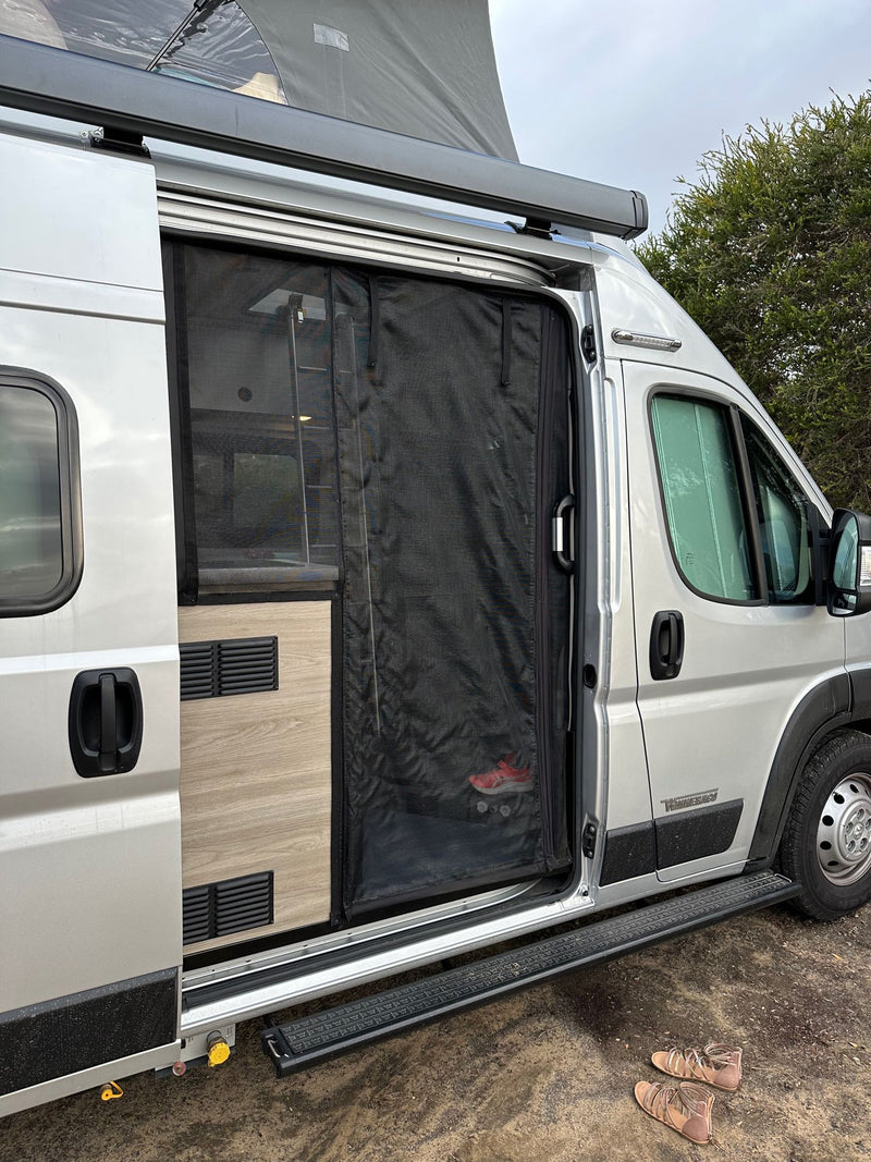  ready for outdoor adventures.|A silver pop-top RV rental is parked on a gravel site amidst tall pine trees in a busy campground.|Open side door of a silver RV rental camper van with mesh screen — photo 3