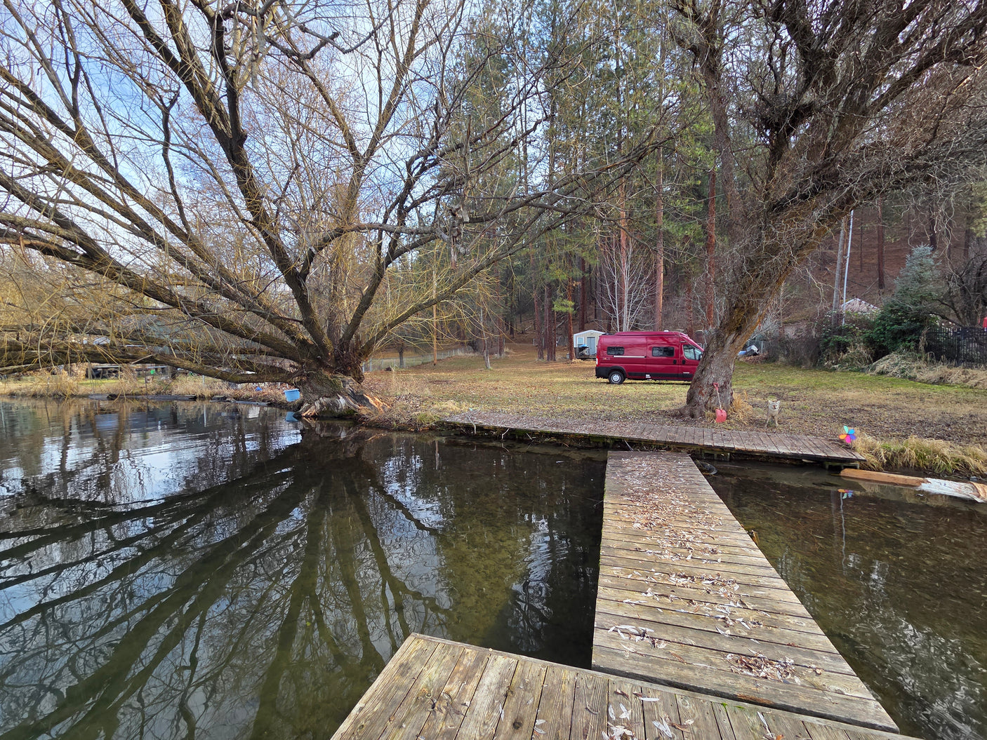 A red RV is parked by a peaceful lakeside dock with bare trees and a coyote. Ideal for a serene nature getaway or an RV rental adventure.