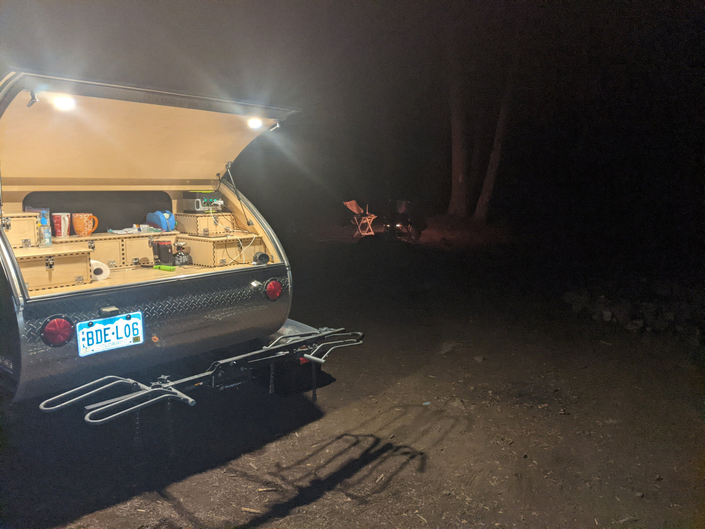 Illuminated rv rental teardrop camper galley at night, with a bike rack attached. A cozy campfire glows in the distant dark. — photo 6