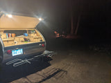 Illuminated rv rental teardrop camper galley at night, with a bike rack attached. A cozy campfire glows in the distant dark.