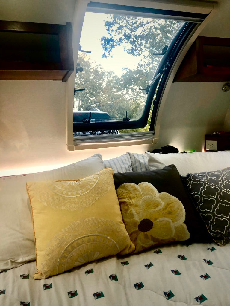 Cozy RV rental interior featuring a bed with decorative pillows and an open window revealing lush trees. #rvrental #camping #travel