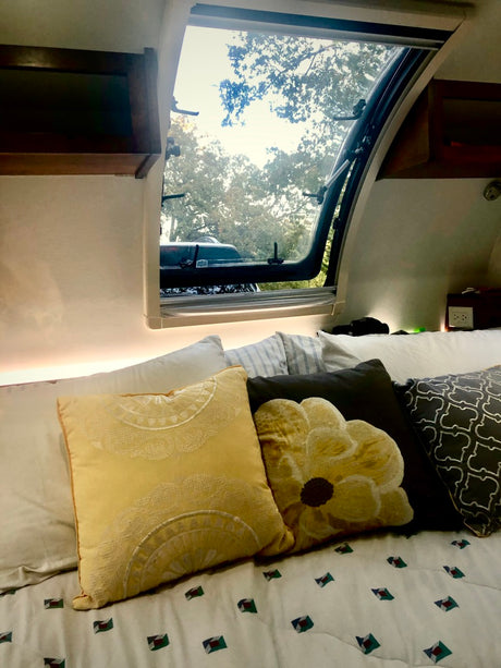 Cozy RV rental interior featuring a bed with decorative pillows and an open window revealing lush trees. #rvrental #camping #travel