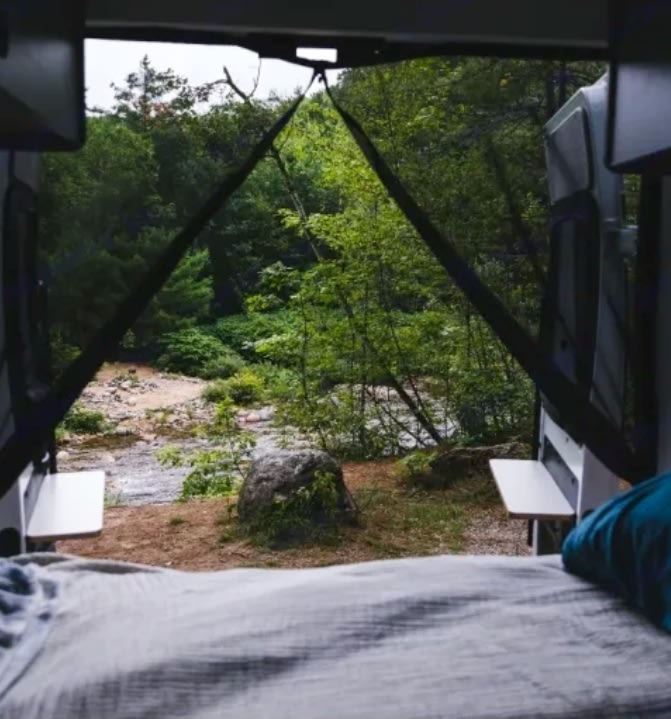 View from an **RV rental** bed looking out at a scenic river, green trees, and a large rock by the water.