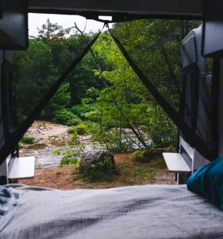 View from an **RV rental** bed looking out at a scenic river, green trees, and a large rock by the water.