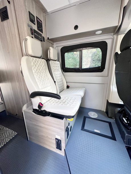 Inside a modern RV rental: white quilted seating, various control panels, an overhead cabinet, and blue patterned flooring.